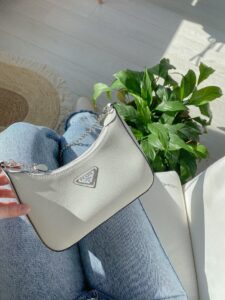 Stylish white designer handbag resting on denim jeans, surrounded by indoor sunlight and plants.